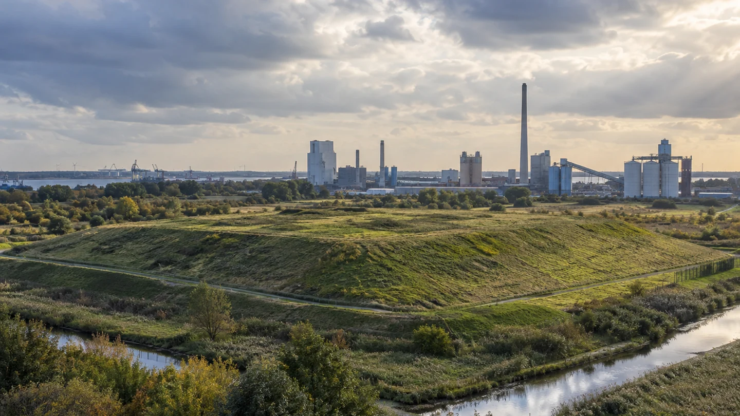 Symbolische Darstellung einer begrünten historischen Industrieablagerung vor Industrieanlagen; KI-Illustration, kein Foto aus Nordenham